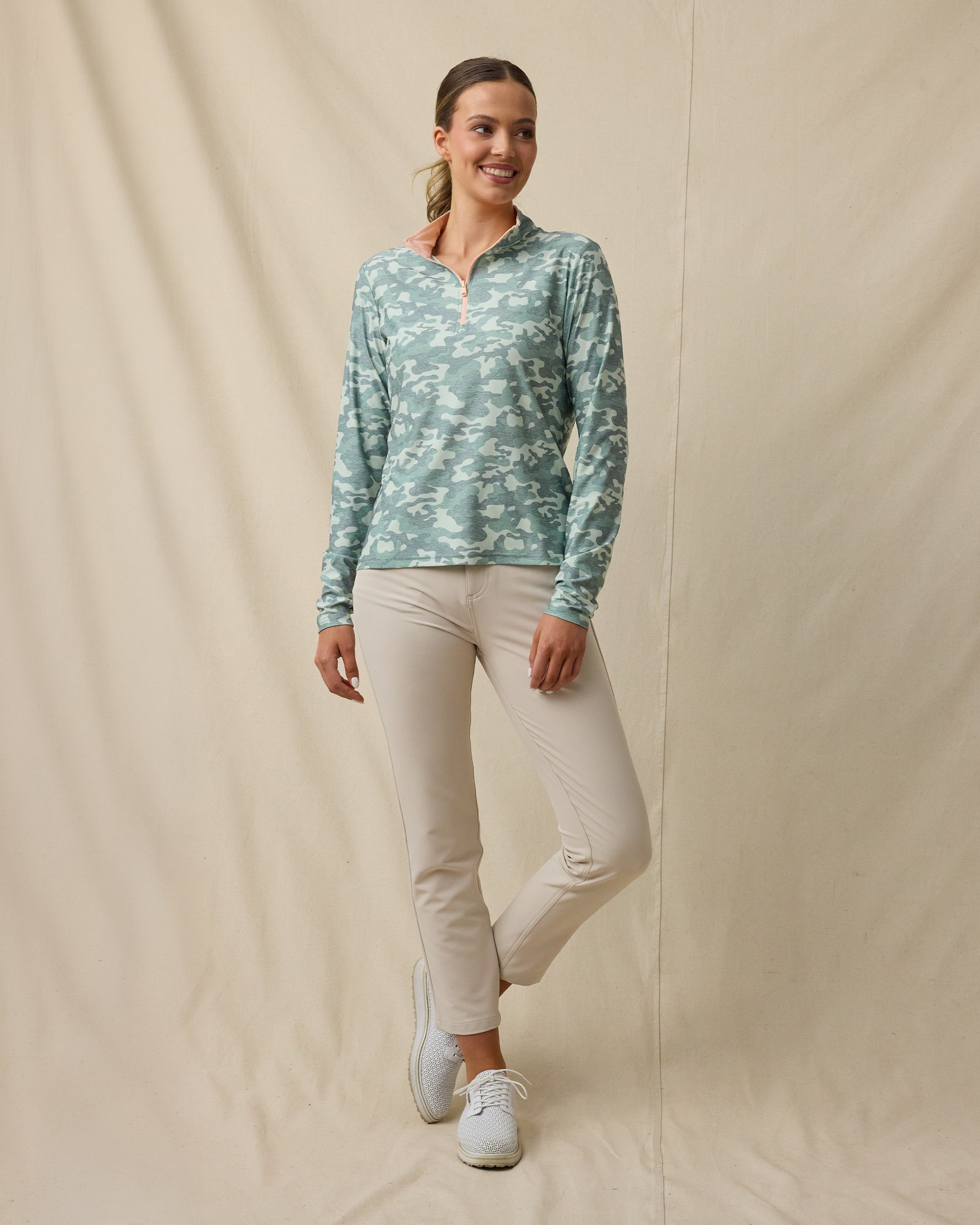 Full-length view of a woman wearing a lighter and darker green camouflage quarter-zip top with a light peach contrast collar, beige pants, and white golf shoes.