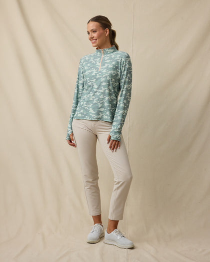 Full-length front view of a woman wearing a green camouflage quarter-zip top zipped to the top, paired with beige pants and white golf shoes.