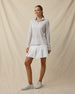 Full-length front view of a woman wearing a light blue pastel floral quarter-zip top with a white pleated skirt and white golf shoes.