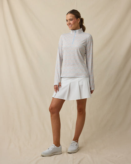 Full-length view of a woman wearing a light blue floral quarter-zip top zipped to the top, paired with a white pleated skirt and white golf shoes.