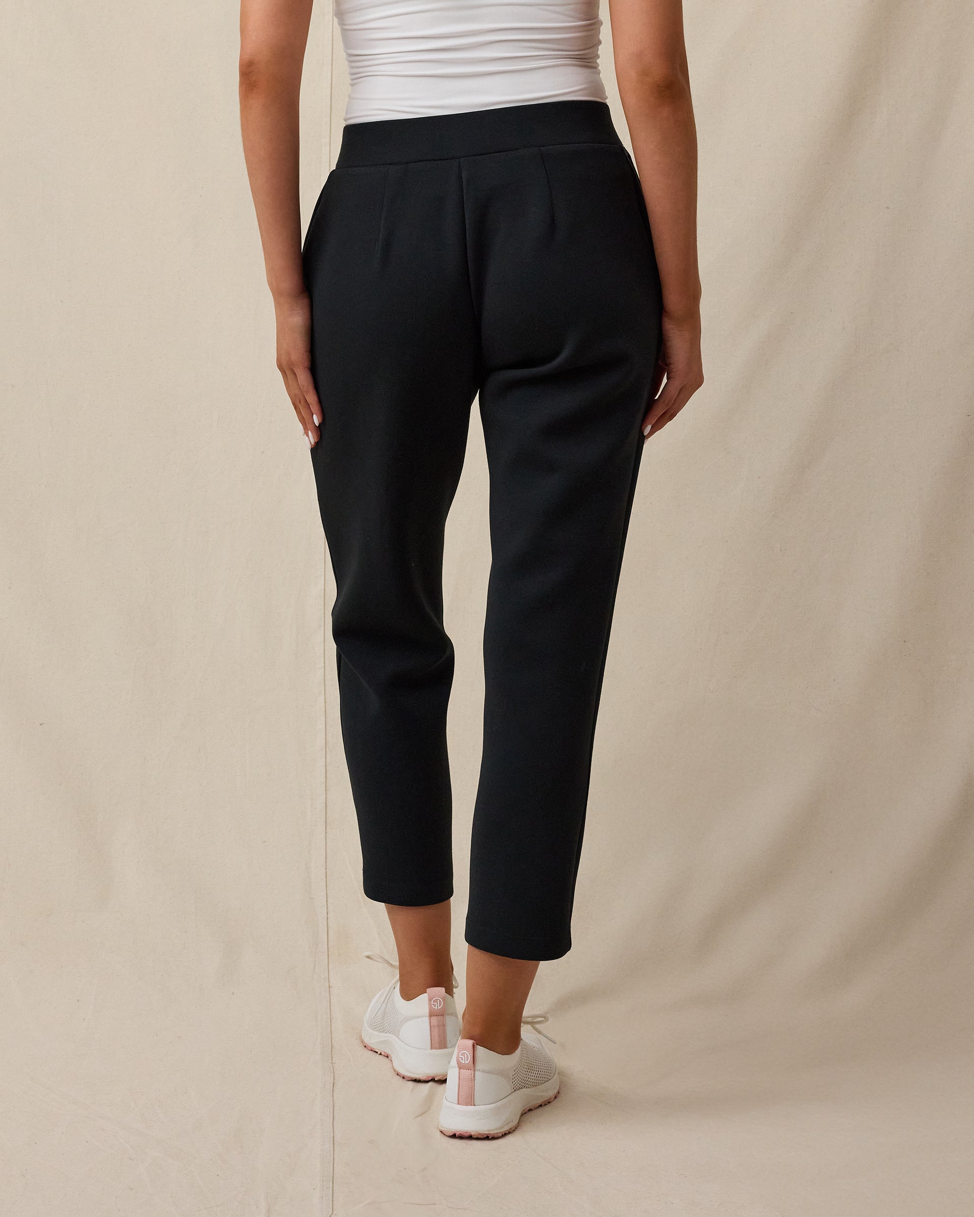 The back view of a women modeling black cropped pants, a white top, and white sneakers against a beige studio backdrop.
