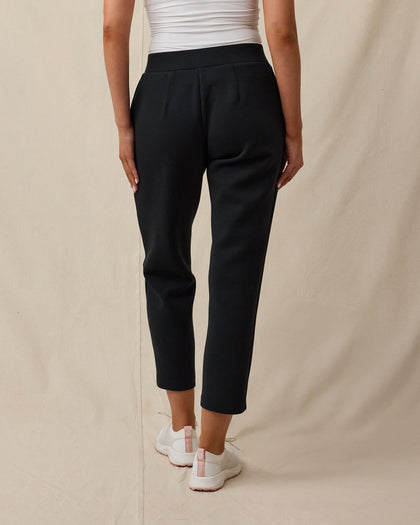 The back view of a women modeling black cropped pants, a white top, and white sneakers against a beige studio backdrop.