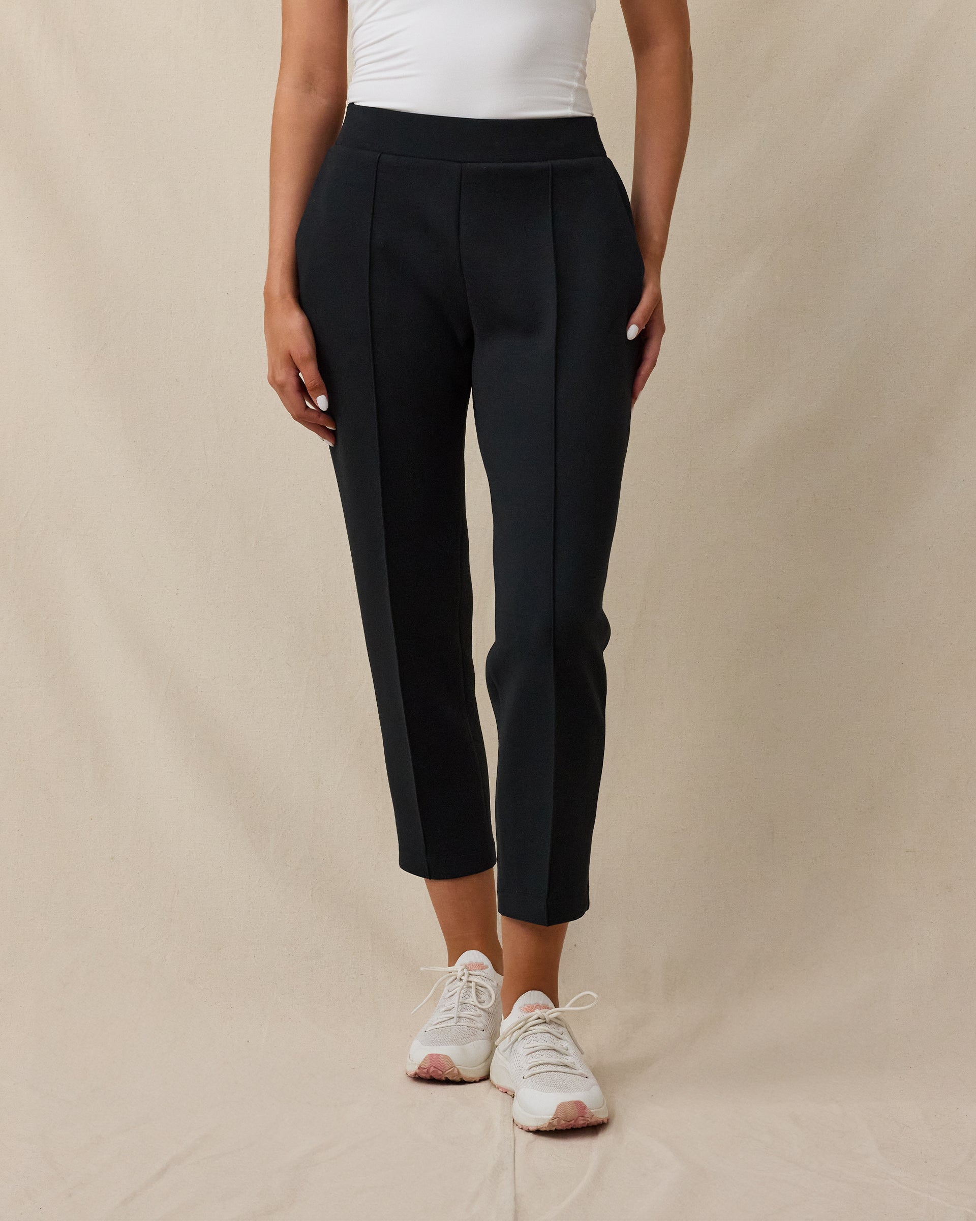 A model wearing black cropped pants with front pleats, paired with a white top and white sneakers, standing against a beige fabric backdrop.