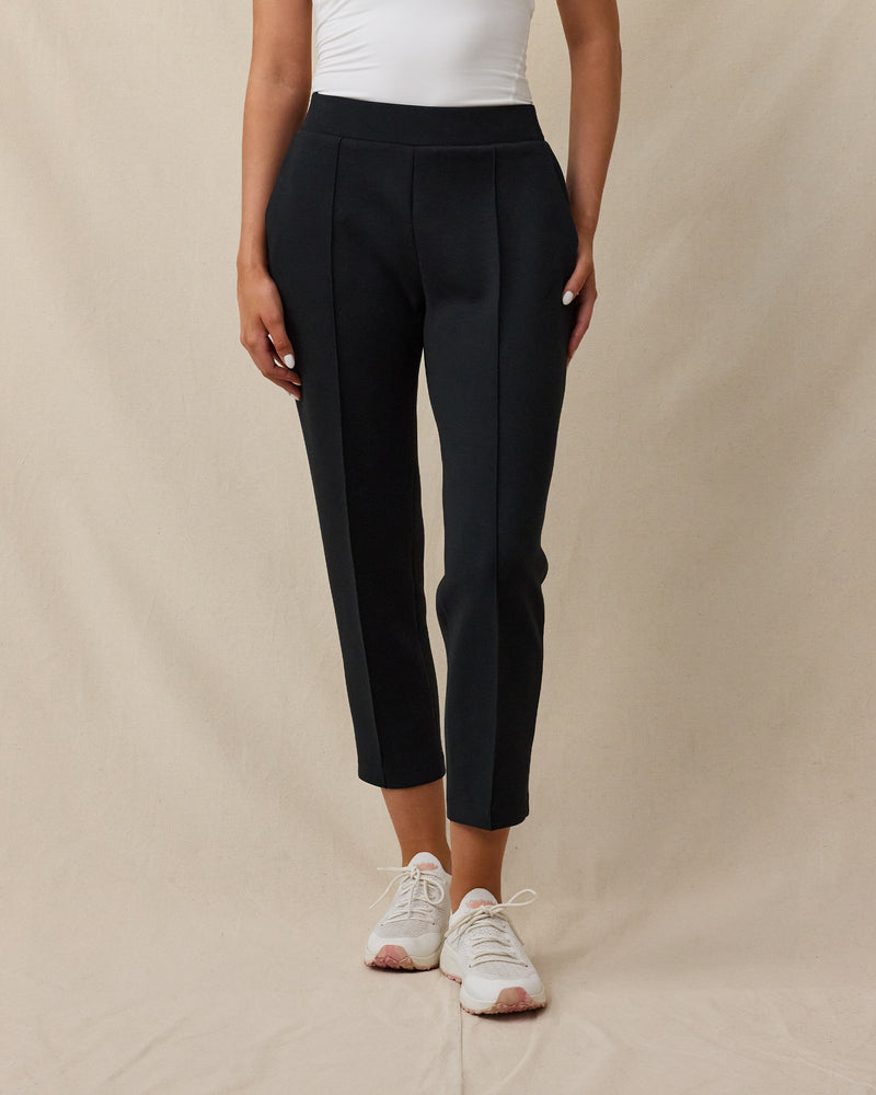 A model wearing black cropped pants with front pleats, paired with a white top and white sneakers, standing against a beige fabric backdrop.