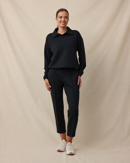 A woman modeling a black long-sleeve polo shirt and matching black cropped trousers, paired with white sneakers, against a draped beige fabric backdrop.