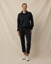 A woman modeling a black long-sleeve polo shirt and matching black cropped trousers, paired with white sneakers, against a draped beige fabric backdrop.