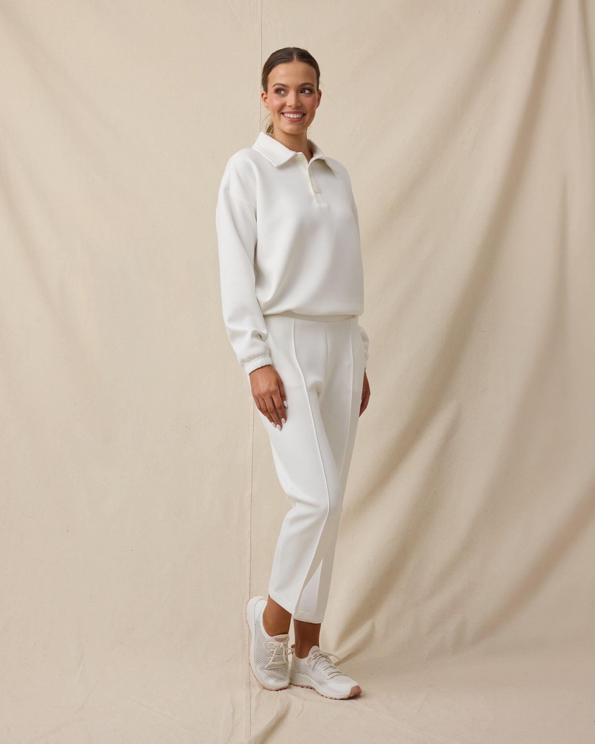 Woman wearing the Chelsea Pant in Coconut with a coordinating off-white long-sleeve top and white sneakers.