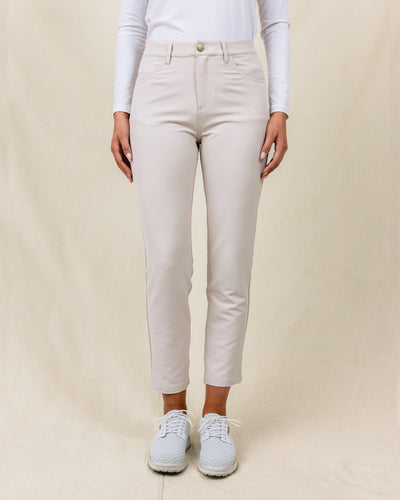 Women's Stride Pant in Stone, front view on model featuring a stone colored ankle pant with front patch pockets and gold button closure, paired with a white long sleeve polo and white golf shoes
