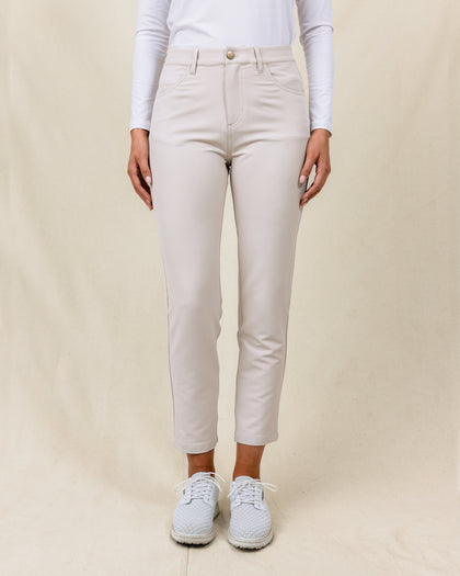 Women's Stride Pant in Stone, front view on model featuring a stone colored ankle pant with front patch pockets and gold button closure, paired with a white long sleeve polo and white golf shoes