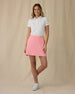 Women's Evie Skort in Geranium, full length front view on model featuring a pink skort with two front pockets, paired with a white polo and white golf shoes