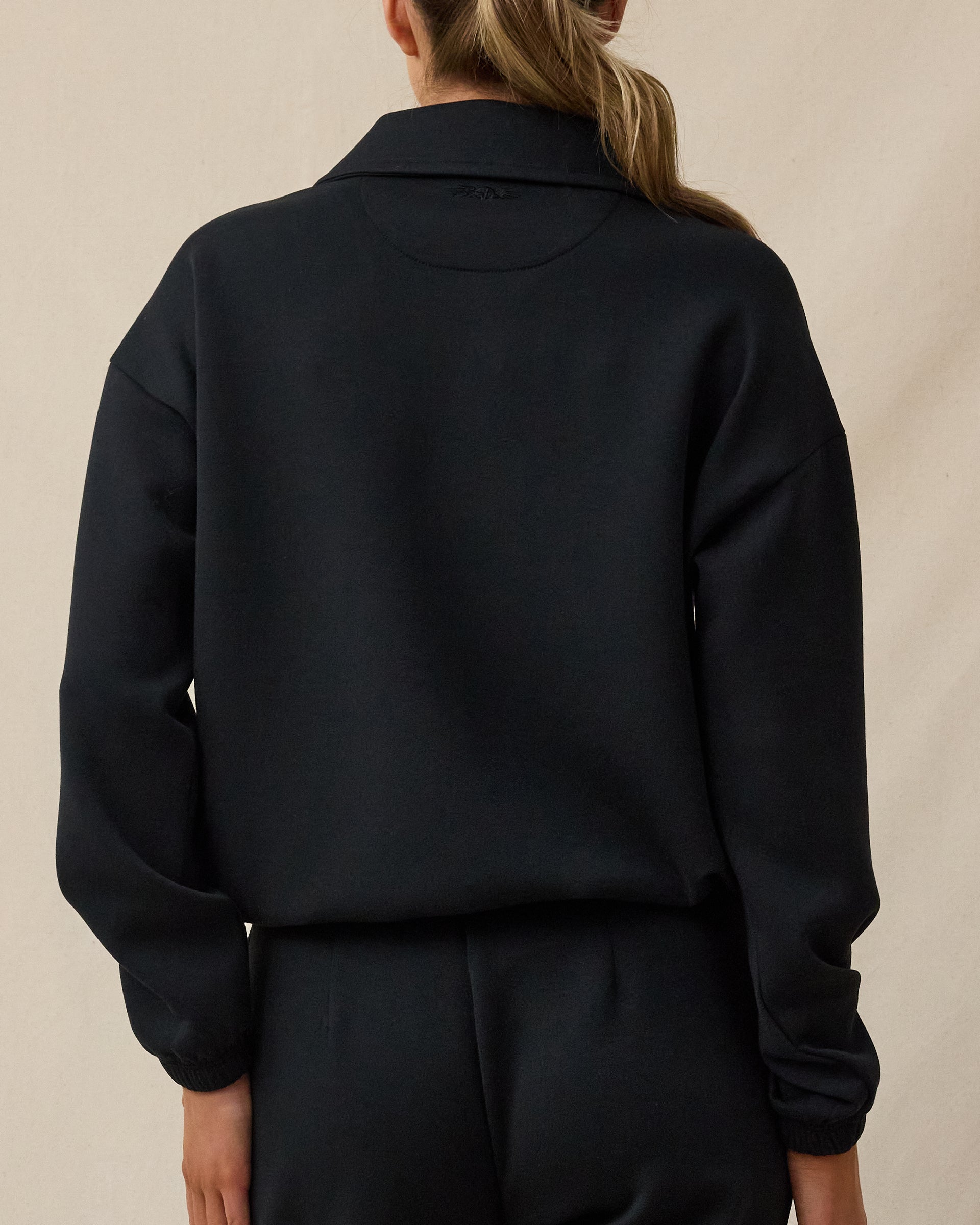 The back view of a woman modeling a black long-sleeve cropped sweatshirt or jacket with a collar, paired with black pants, against a plain beige studio background.