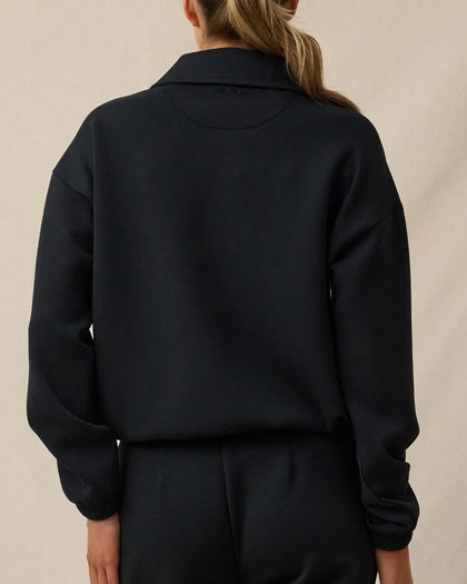 The back view of a woman modeling a black long-sleeve cropped sweatshirt or jacket with a collar, paired with black pants, against a plain beige studio background.