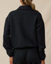 The back view of a woman modeling a black long-sleeve cropped sweatshirt or jacket with a collar, paired with black pants, against a plain beige studio background.