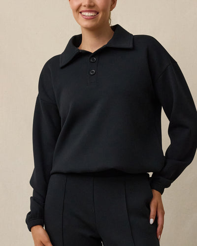 A woman modeling a black long-sleeve polo sweater with a collar and two-button placket, paired with black pants, against a plain beige background.