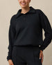 A woman modeling a black long-sleeve polo sweater with a collar and two-button placket, paired with black pants, against a plain beige background.