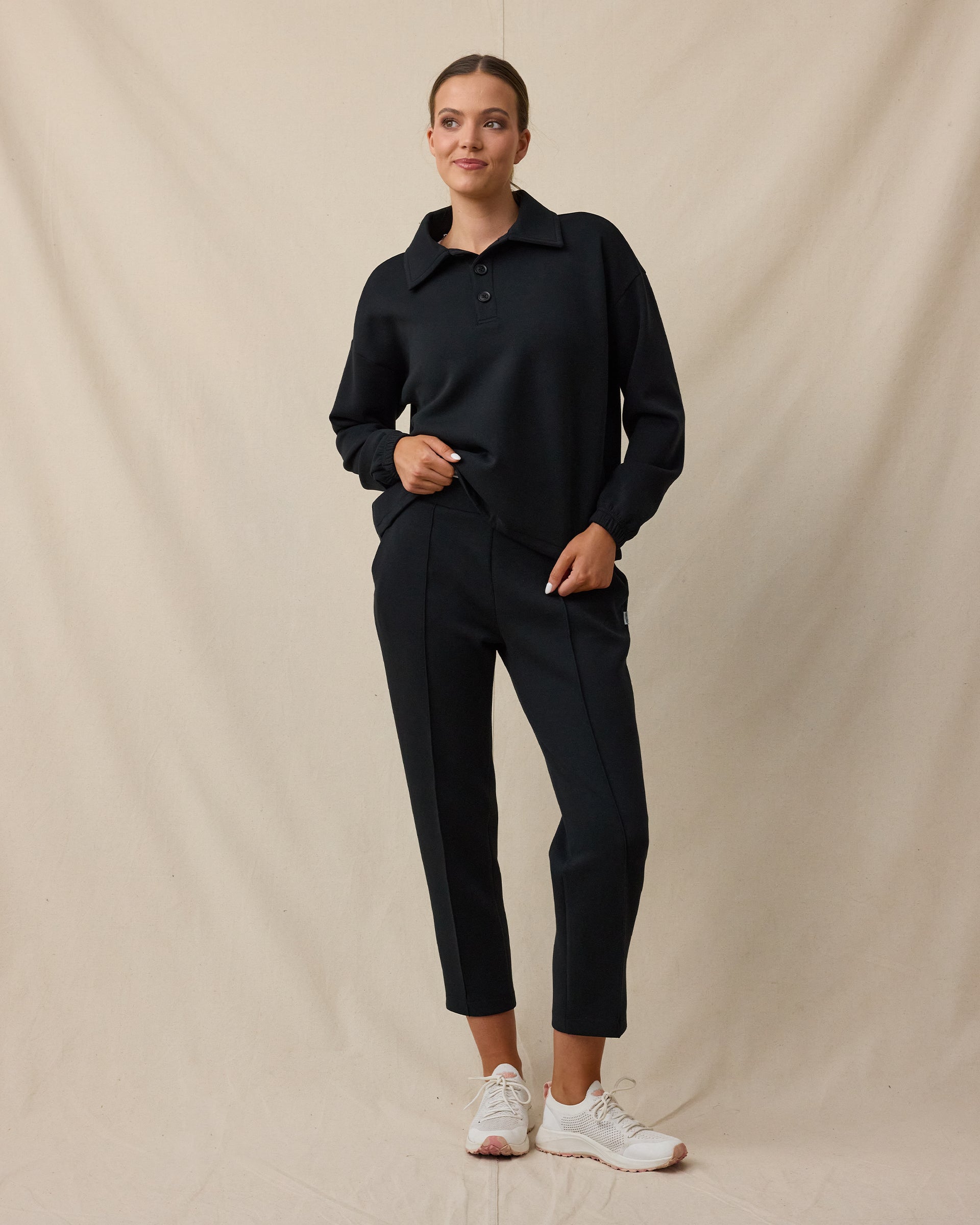 A woman modeling a black long-sleeve collared top and matching black cropped pants, paired with white sneakers, standing against a draped beige fabric backdrop.