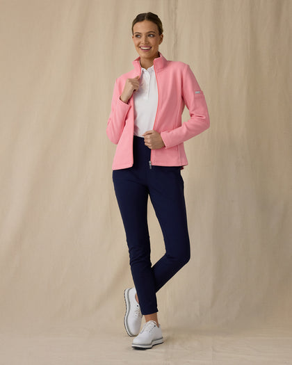 Women's Swing Jacket in Geranium, full length front view on model featuring a pink full zip jacket with a ribbed texture worn open over a white polo, paired with navy pants and white golf shoes