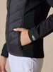 Nova Jacket in Black, close-up front view on model featuring the diagonal quilted body, side zip pocket with hand inserted, and silver embroidered logo detail on the cuff styled with white pants