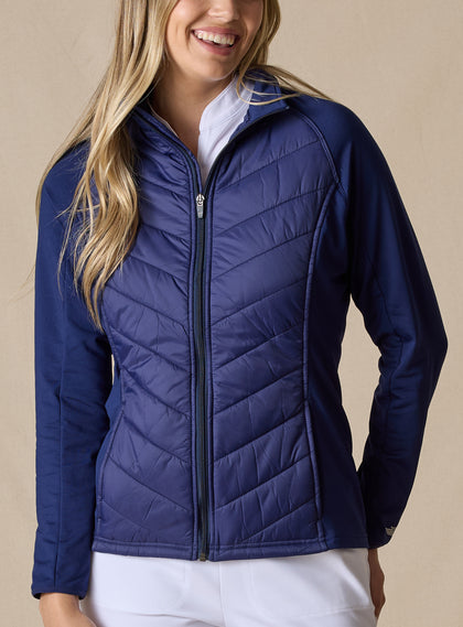 Nova Jacket in Indigo, front view on model featuring a navy hybrid full-zip jacket with quilted body, stretch knit sleeves, and mock neck collar styled with white pants