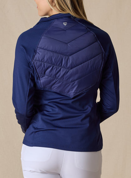 Nova Jacket in Indigo, back view on model featuring a navy hybrid jacket with diagonal quilted upper back panel, stretch knit lower back and sleeves, and tonal logo detail at the center back neck styled with white pants