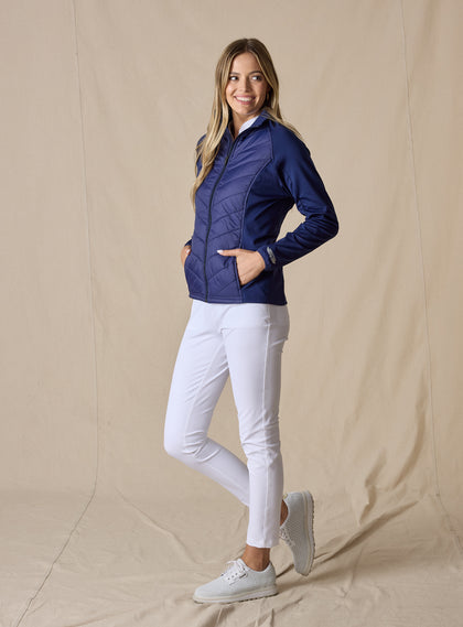 Nova Jacket in Indigo, side view on model featuring a navy hybrid full-zip jacket with diagonal quilted front, stretch knit sleeves, and side zip pockets styled with white pants and white shoes
