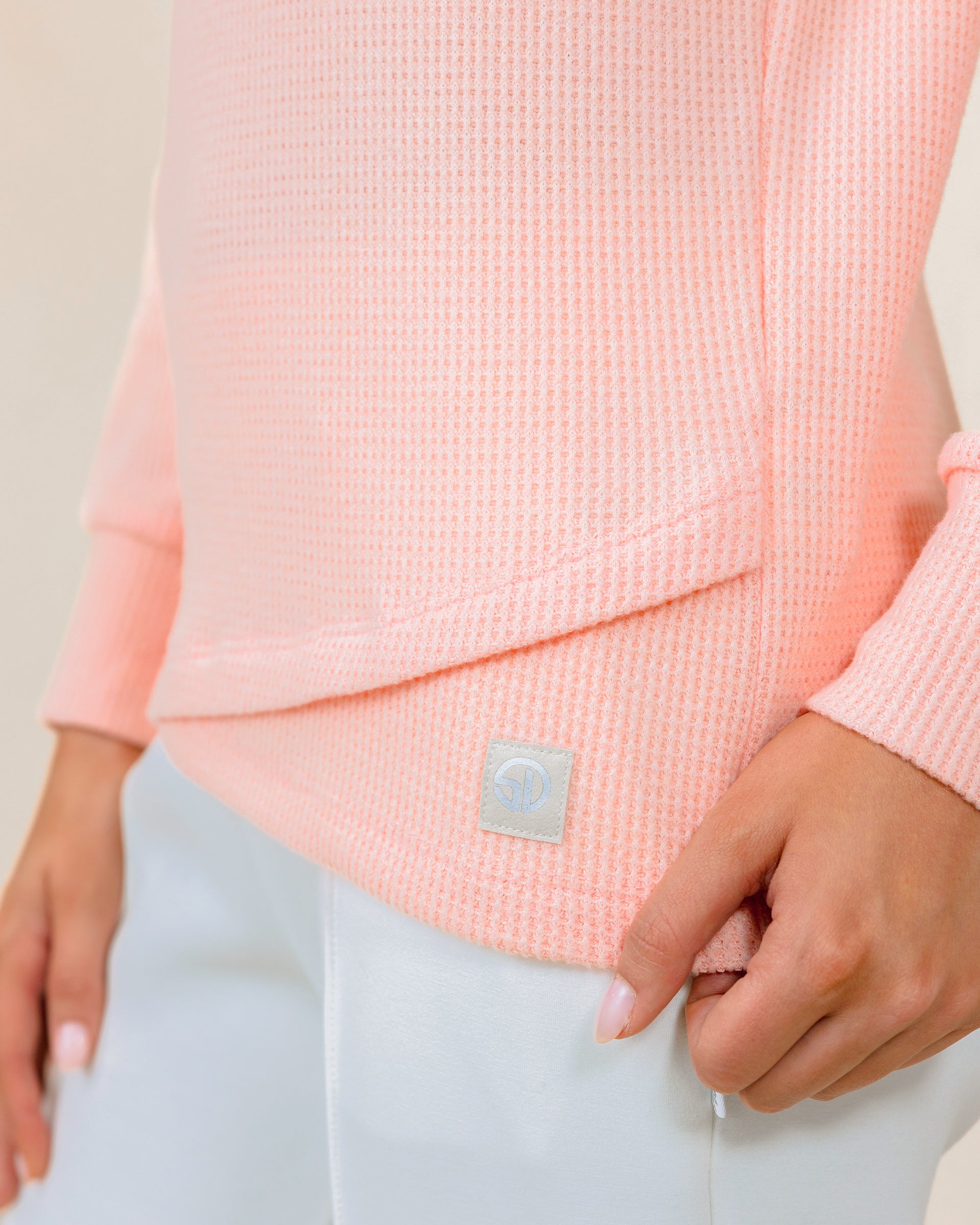 A close-up of a model wearing a soft peach waffle-knit long-sleeve pullover and white pants, with a small square brand patch near the hem.