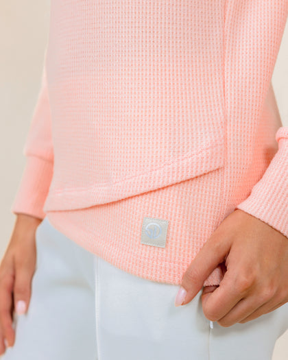 A close-up of a model wearing a soft peach waffle-knit long-sleeve pullover and white pants, with a small square brand patch near the hem.