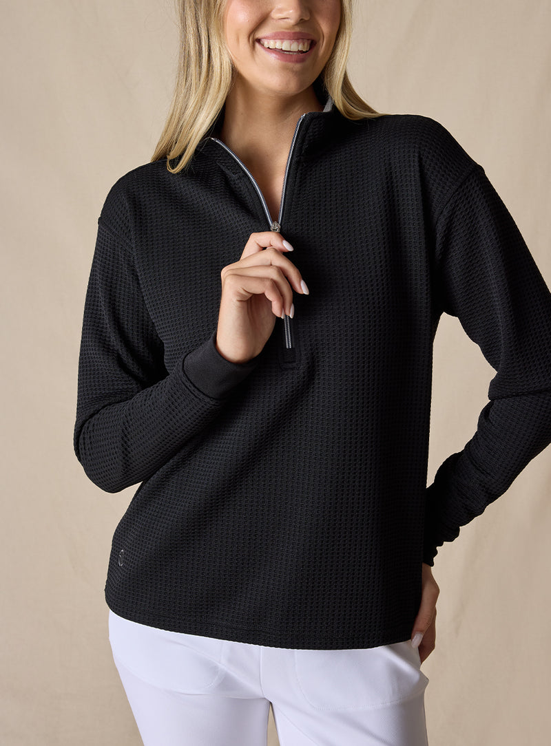 Women's Opal Half Zip in Black, front view on model featuring a black waffle-textured pullover with a quarter-zip neckline and contrast striped zipper paired with white pants