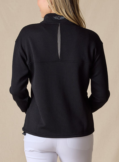 Women's Opal Half Zip in Black, back view on model featuring a black waffle-textured pullover with an embroidered logo at the collar and a center back vent detail paired with white pants