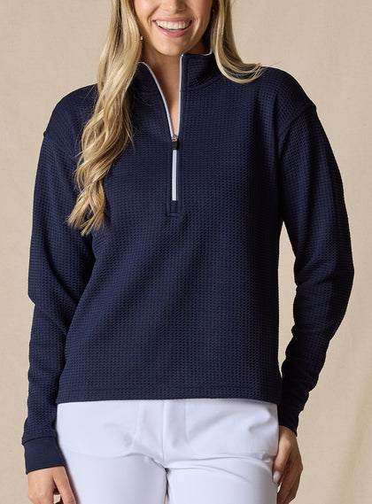 Women's Opal Half Zip in Indigo, front view on model featuring a navy waffle-textured pullover with a quarter-zip neckline and contrast white zipper paired with white pants