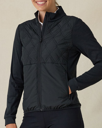 Women's Hera Jacket in Black, front view on model featuring a diamond quilted front body with solid black knit sleeves, full-zip stand collar, and zippered side pocket detail, layered over a white polo and paired with black pants