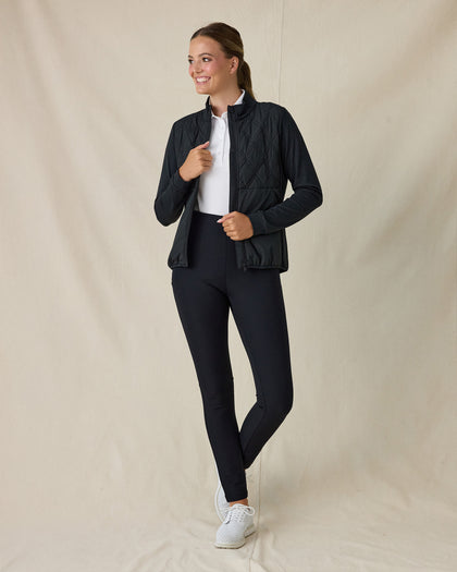 Women's Hera Jacket in Black, full-length front view on model featuring a diamond quilted front body with solid black knit sleeves and full-zip stand collar, worn open and layered over a white polo, paired with black pants and white shoes