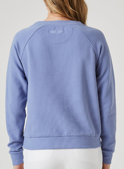 Delia Sweatshirt in Blueberry, back view on model featuring a medium blue ribbed-texture crewneck pullover with raglan sleeves and white embroidered logo detail at the center back neck styled with white pants