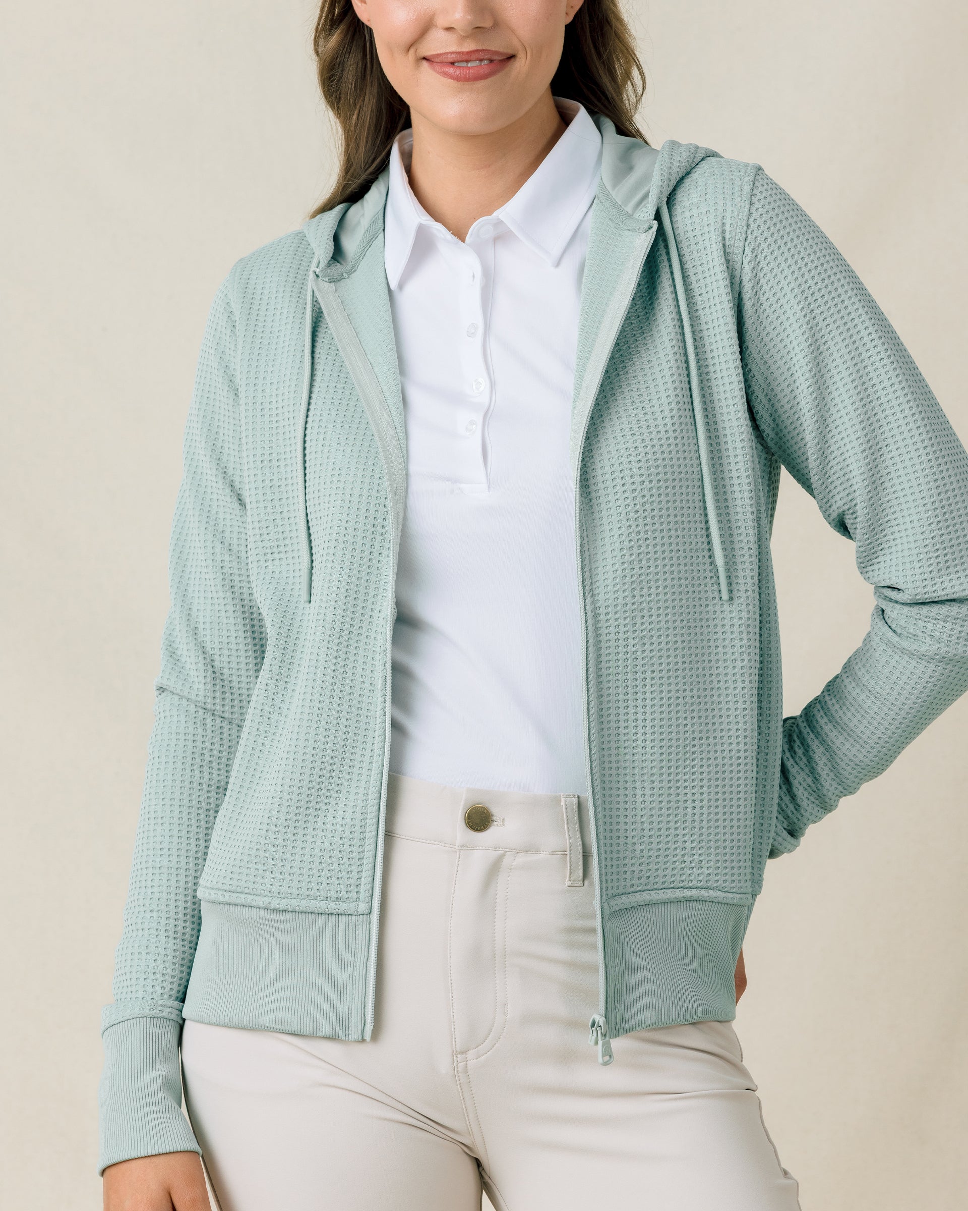 Front view of a model wearing the Eliza Hoodie in Herb open over a white polo with light-colored pants.