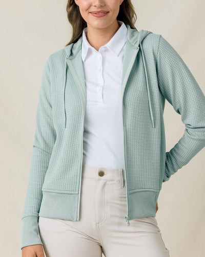 Front view of a model wearing the Eliza Hoodie in Herb open over a white polo with light-colored pants.
