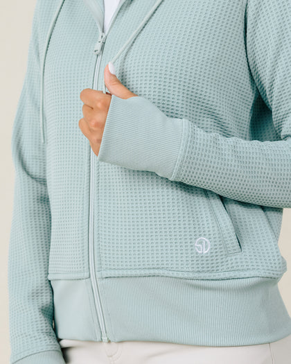 Close-up of the Eliza Hoodie in Herb highlighting the waffle knit texture, thumbhole cuff, front pocket, ribbed hem, and embroidered logo.