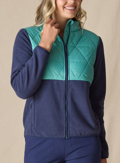 Women's Laurel Hybrid Fleece Jacket in Sea Green and New Indigo, front view on model featuring a sea green quilted upper body with navy fleece lower body and sleeves, full-zip closure, and side zip pocket paired with navy pants