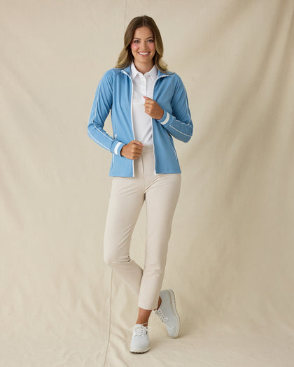 Patty Jacket in Coastal, full length front view on model featuring a solid medium blue full zip jacket with white piping along the sleeves and white striped cuffs, layered over a white polo and paired with khaki ankle pants and white golf shoes