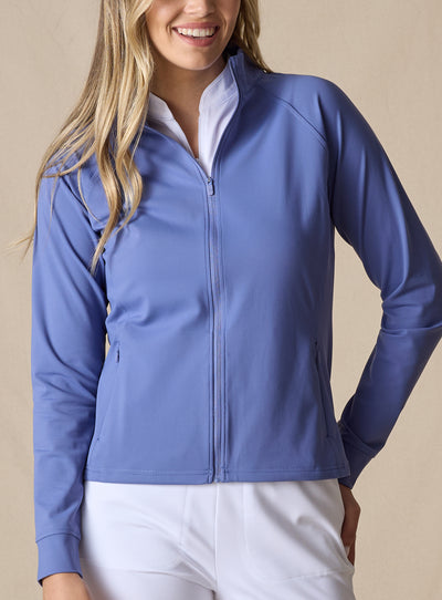 Women's Eirene Jacket in Blueberry, front view on model featuring a solid cornflower blue performance fabric with a full-zip stand collar and raglan sleeves, with zippered side pocket detail, layered over a white polo and paired with white pants