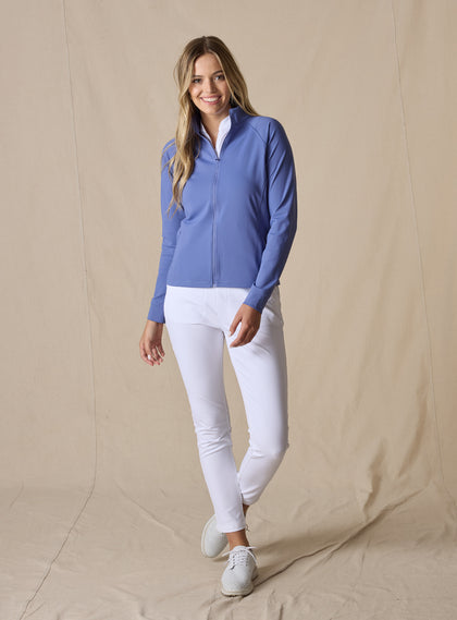 Women's Eirene Jacket in Blueberry, full-length front view on model featuring a solid cornflower blue performance fabric with a full-zip stand collar and raglan sleeves, layered over a white polo and paired with white pants and white shoes