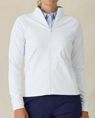 Women's Eirene Jacket in White, front view on model featuring a clean white performance fabric with a full-zip stand collar, raglan sleeves, and zippered side pockets, layered over a light blue polo and paired with navy pants

