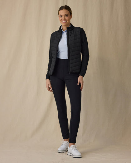 Women's Ida Hybrid Jacket in Black, full length front view on model featuring a black full zip hybrid jacket worn open over a light blue polo, paired with black pants and white golf shoes