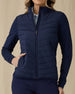 Women's Ida Hybrid Jacket in New Indigo, front close-up view featuring a full-zip closure, horizontal quilted chest and front body, stretch side panels, and hand in side pocket worn over a light blue polo