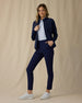 Women's Ida Hybrid Jacket in New Indigo, front full-length view featuring an open zip, horizontal quilted front body, stretch side panels, and side pocket with hand inserted paired with navy pants and white shoes