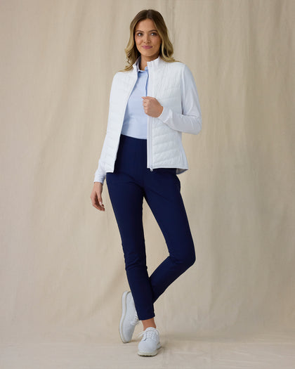 Women's Ida Hybrid Jacket in White, front full-length view featuring an open zip, horizontal quilted front body, and stretch side panels paired with navy pants and white shoes worn over a light blue polo