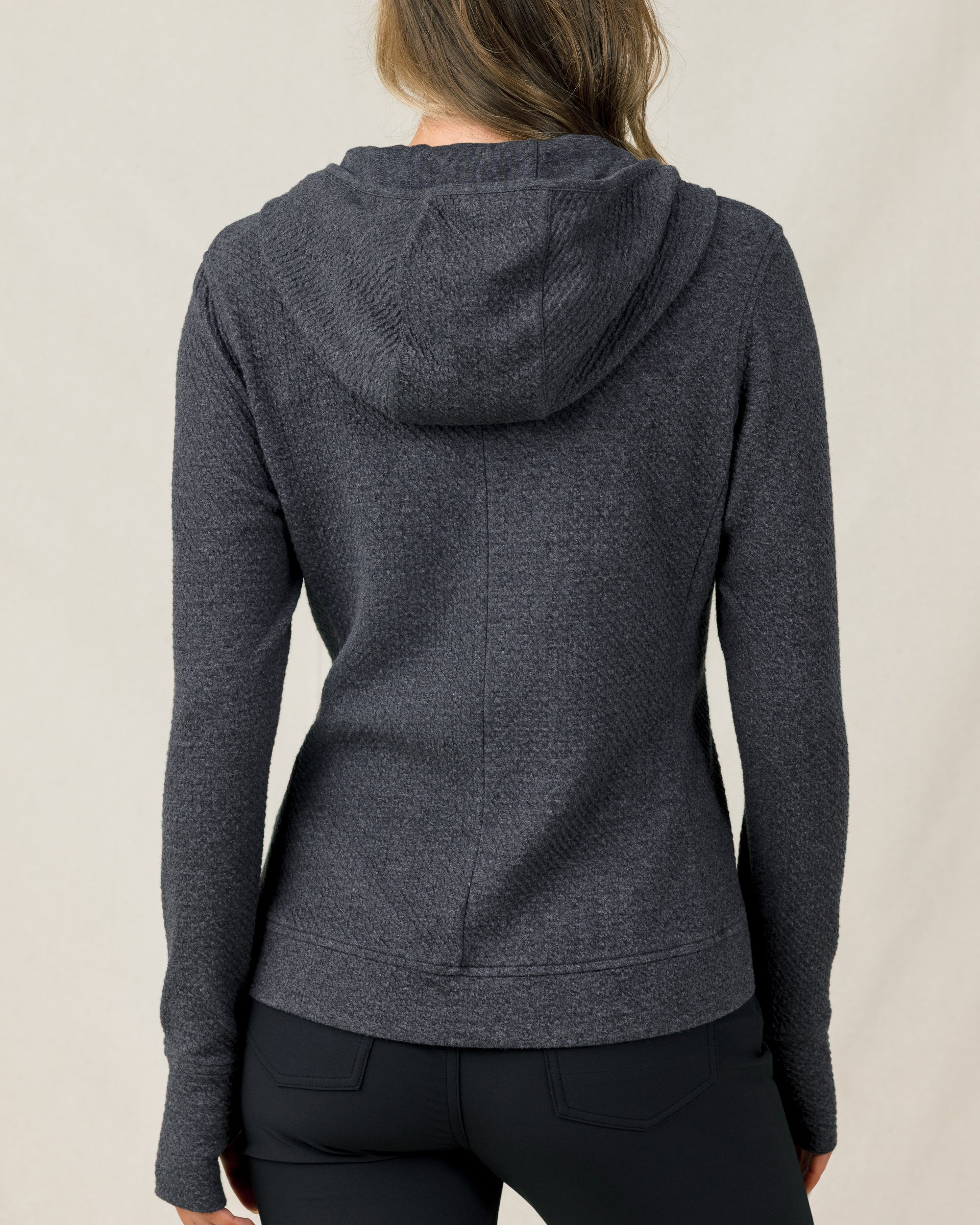 The back view of a woman modeling a dark heather gray textured hooded sweater and black pants, against a plain light beige studio background.