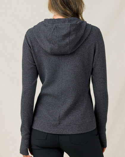 The back view of a woman modeling a dark heather gray textured hooded sweater and black pants, against a plain light beige studio background.