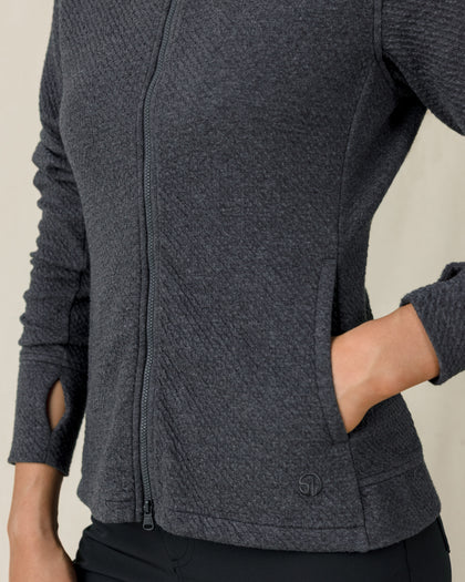 Close-up of a charcoal gray textured zip-up jacket showing the front zipper, hand pocket, thumbhole cuff, and subtle logo near the hem.