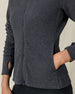 Close-up of a charcoal gray textured zip-up jacket showing the front zipper, hand pocket, thumbhole cuff, and subtle logo near the hem.