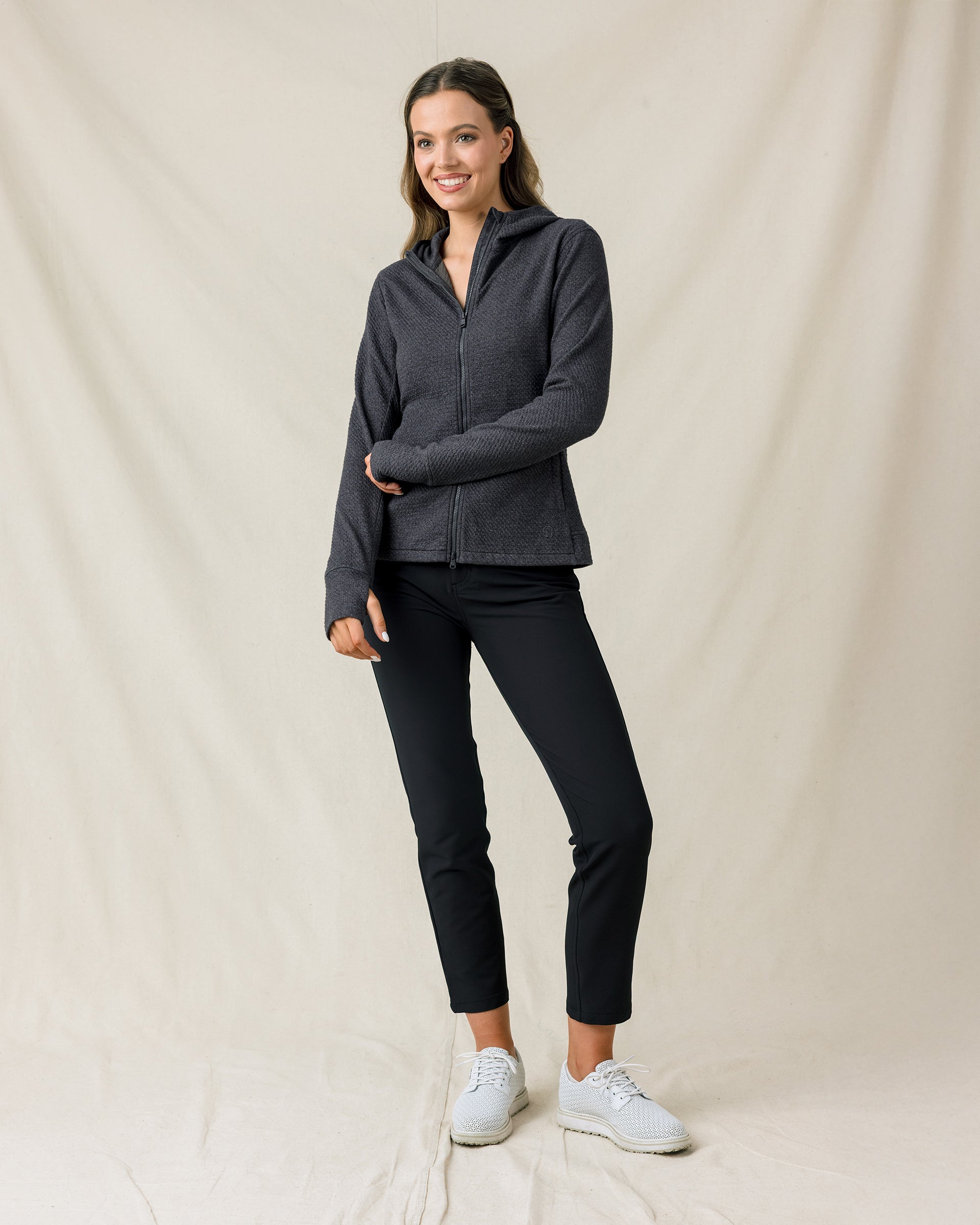 A woman modeling a dark gray zip-up athletic jacket and black cropped pants with white sneakers, standing against a draped beige fabric backdrop.